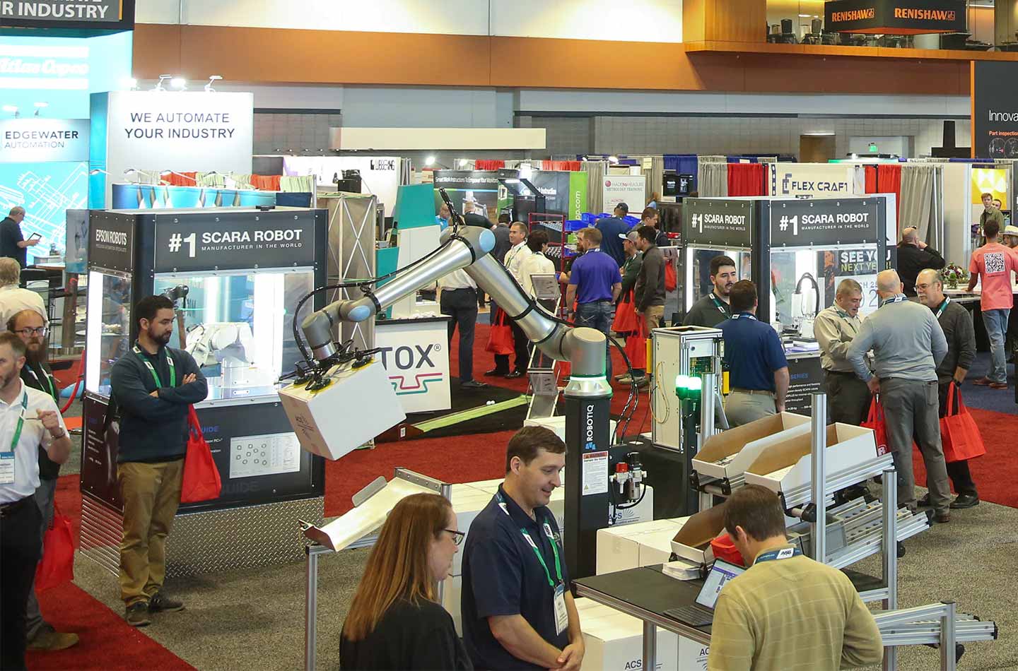 Trade show floor featuring multiple automation and robotics booths, including a robotic arm lifting a box in the foreground