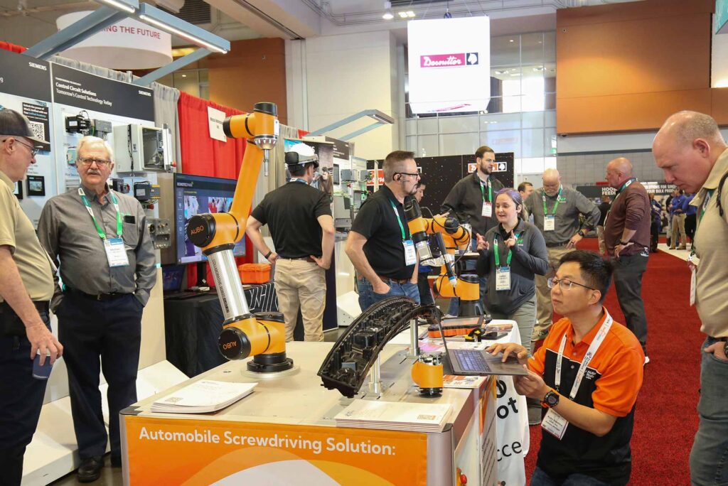 People interacting at an industrial automation trade show booth featuring robotic arms, with multiple conversations and demonstrations happening simultaneously